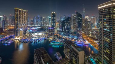 Dubai Marina hava saatinin gece aydınlanması, BAE. Modern gökdelenler ve konut binaları. Yollarda trafik vardı. Yapay kanal şehrindeki alışveriş merkezinin yakınındaki yatlar ve tekneler.