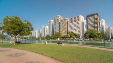 Abu Dabi 'nin sahil şeridi boyunca uzanan Corniche Bulvarı plajı parkında arka planda gökdelenler var. Lake Park 'ın manzarası. Bir tarafta ağaçlar. Güneşli bir günde mavi gökyüzü