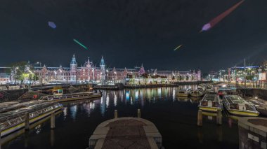 Panorama aydınlatılmış Central Railway İstasyonu gece saat çizelgesini gösteriyor. Hollanda, Amsterdam 'daki tren istasyonunun önündeki bir iskeleye, tramvaylara ve kanallara demirlemiş tur tekneleri