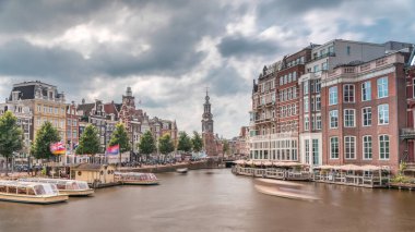 Amstel Nehri, rıhtım ve Munttoren kulesinin yer aldığı Amsterdam şehir merkezinin panoramik zaman atlaması. Tekneler bulutlu gökyüzünün altında ileri geri yüzerler. Köprüden manzaralı şehir manzarası. Hollanda