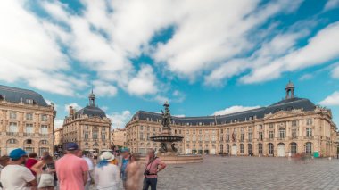 Place de la Bourse zaman atlaması Fransa 'nın Bordeaux şehrinde, Bulutlu mavi gökyüzünün altında klasik mimari ile çevrili Üç Zarafet Çeşmesi ile birlikte. Meydanda yürüyen insanlar.
