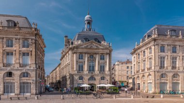 Place de la Bourse, Fransa 'nın Bordeaux şehrinde, bulutlu mavi gökyüzü altında klasik mimari ile çevrili restoranları olan bir yer. Meydanda ve ana caddelerde yürüyen insanlar
