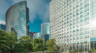 Panorama modern ofis binalarının zaman çizelgesini gösteriyor. Parktaki yeşil ağaçlar tarafından çerçevelenmiş parlak mavi gökyüzü üzerinde yansımaları ve röfleleri olan cam gökdelenler. Brüksel 'de güneş ışığı ve geçen bulutlar