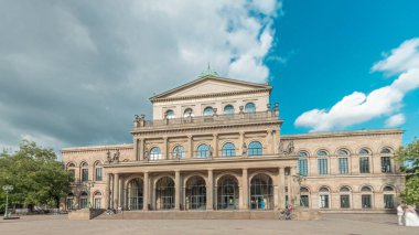 Hanover Devlet Operası 'nın (Staatsoper Hannover) ön görüntüsü. Şehir meydanında canlı mavi bulutlu bir zaman dilimi altında yürüyen insanların olduğu klasik tarzda bir tiyatro. Almanya