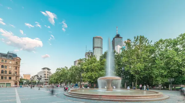 Frankfurt, Almanya merkezindeki Opera Meydanı (Opera Meydanı) zaman atlaması, arka planda gökdelenler ve kulelerle çevrili ve akşam gökyüzü altında ağaçlarla çevrili bir çeşme ve şehir hayatı sergiliyor.