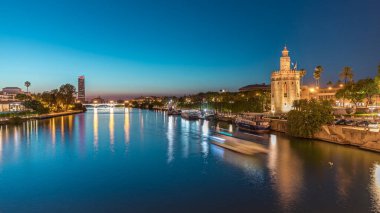 İspanya, Seville 'deki Torre del Oro gözlem kulesi yakınlarındaki Guadalquivir Nehri boyunca yüzen bir tur teknesi. Günbegün geçiş zamanı, şehrin ışıkları su ve deniz manzarasını yansıtıyor.