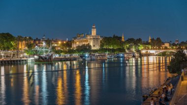 İspanya, Sevilla 'daki Torre del Oro gözlem kulesi. Günbegün geçiş zamanı, şehrin ışıkları su ve deniz manzarasını yansıtıyor. Guadalquivir Nehri boyunca yüzen bir tekne.