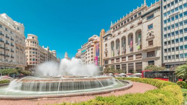 Panorama, İspanya 'nın Valencia şehrinde Belediye Meydanı' nda (Plaza del Ayuntamiento Meydanı) fıskiyeyi gösteriyor. Büyük mimari, çiçek açan yeşillik ve mavi gökyüzünün altında su jetleri. İnsanlar meydanda geziniyor.