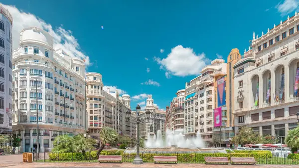 Valencia, İspanya 'da Belediye Meydanı' ndaki (Plaza del Ayuntamiento Meydanı) zaman atlaması. Büyük mimari, çiçek açan yeşillik ve bulutlu bir gökyüzünün altında su jetleri. İnsanlar canlı meydanda geziniyor..