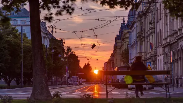 Street with sunset in the Croatian capital Zagreb. People sitting on the bench near tree. Road traffic and tram rails