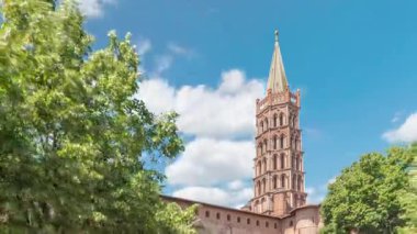 Fransa 'nın Toulouse kentindeki Saint Sernin Bazilikası' nın kaldırımlı parvisleri. Zaman atlaması. Avrupa 'nın en büyük Roma kilisesi tuğla çan kulesi, kemerli kapılar, yeşil ağaçlar ve mavi bulutlu gökyüzü.