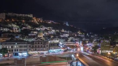Gürcistan 'ın Tiflis şehrinde ahşap oyulmuş balkonlu renkli geleneksel evleri gösteren panorama. Aydınlatılmış binalar ve nehir üzerindeki bir köprüde trafik