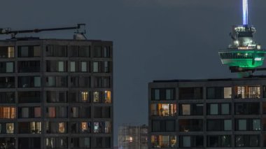 Rotterdam gece şehir manzarası. Euromast gözlem kulesi ve gökdelenlerin parlayan pencereleri var. Rıhtımda modern gökdelenler ve manzaralı bir liman manzarası yer alıyor.