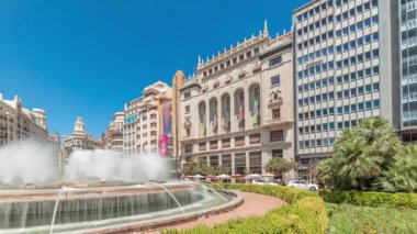 Panorama, Valencia, İspanya 'da Belediye Meydanı Plaza del Ayuntamiento zaman kapsülünü gösteriyor. Büyük mimari, çiçek açan yeşillik ve mavi gökyüzünün altında su jetleri. İnsanlar meydanda geziniyor.