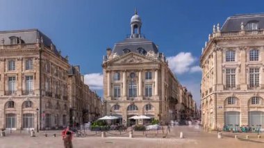 Place de la Bourse, Fransa 'nın Bordeaux şehrinde, bulutlu mavi gökyüzü altında klasik mimari ile çevrili restoranları olan bir yer. Meydanda ve ana caddelerde yürüyen insanlar