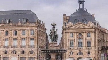 Place de la Bourse zaman atlaması Fransa 'nın Bordeaux şehrinde, Bulutlu mavi gökyüzünün altında klasik mimari ile çevrili Üç Zarafet Çeşmesi ile birlikte. İnsanlar ana meydanda bir tramvay durağında
