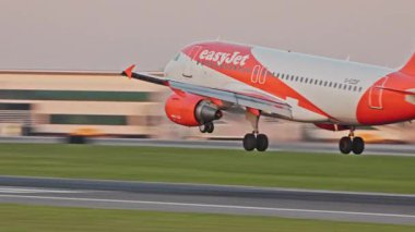 Bir ticari uçak EasyJet gün batımında Lizbon Havaalanı 'nın pistine iner ve lastik dumanı yere iner. Terminaller ve açık bir gökyüzü bu dinamik havacılık sahnesinde canlı bir zemin oluşturur.