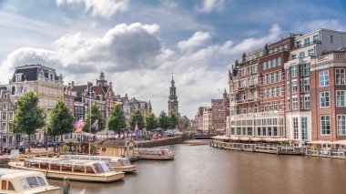 Amstel Nehri, rıhtım ve Munttoren kulesinin yer aldığı Amsterdam şehir merkezinin panoramik zaman atlaması. Tekneler bulutlu gökyüzünün altında ileri geri yüzerler. Köprüden manzaralı şehir manzarası. Hollanda