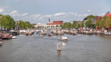 Amstel Nehri 'ndeki Amstel kilitlerinden geçen teknelerin panoramik görüntüsü. Opera binası ve Zuiderkerk kulesi arka planda görünüyor. Bulutlar gökyüzünü dolduruyor. Amsterdam, Hollanda