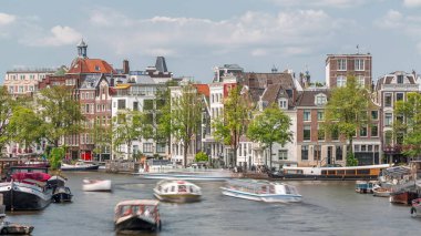 Güneşli bir günde Amsterdam 'ın şehir merkezindeki işlek bir nehrin zamanı. Rıhtımı çevreleyen yeşil ağaçlı tekneler trafiği. Arka planda tipik tarihi binalar. Amsterdam, Hollanda