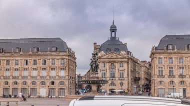 Place de la Bourse zaman atlaması Fransa 'nın Bordeaux şehrinde, Bulutlu mavi gökyüzünün altında klasik mimari ile çevrili Üç Zarafet Çeşmesi ile birlikte. İnsanlar ana meydanda bir tramvay durağında