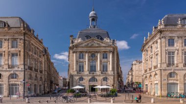 Place de la Bourse, Fransa 'nın Bordeaux şehrinde, bulutlu mavi gökyüzü altında klasik mimari ile çevrili restoranları olan bir yer. Meydanda ve ana caddelerde yürüyen insanlar