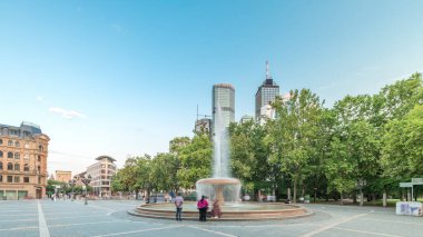 Frankfurt, Almanya merkezindeki Opera Meydanı (Opera Meydanı) zaman atlaması, arka planda gökdelenler ve kulelerle çevrili ve akşam gökyüzü altında ağaçlarla çevrili bir çeşme ve şehir hayatı sergiliyor.