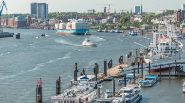 Almanya 'nın Hamburg kentinde Elbe nehir kenarının hava zaman çizelgesi. İskeleye yakın tekneler, Jan Fedder gezinti güvertesi, marina ve konteyner gemilerinin geçtiği manzara. Elbe Nehri kıyısındaki canlı şehir manzarası