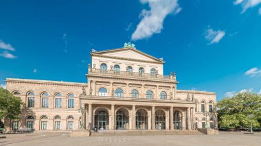Hanover Devlet Operası 'nın (Staatsoper Hannover) ön görüntüsü. Şehir meydanında canlı mavi bulutlu bir zaman dilimi altında yürüyen insanların olduğu klasik tarzda bir tiyatro. Almanya