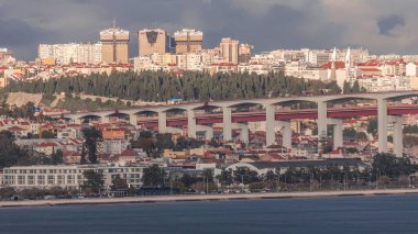 Renkli evler, Amoreiras alışveriş merkezi kuleleri ve 25 Nisan Köprüsü 'nün yer aldığı Lizbon siluetinin hava zaman çizelgesi. Önünde yeşil ağaçlar ve Prazeres Mezarlığı bulunan tarihi bölge