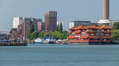 Euromast gözlem kulesi yakınlarındaki Rotterdam şehir manzarası zaman çizelgesi ve Nieuwe Maas Nehri 'ndeki bir turist teknesi. Rıhtımda modern gökdelenler, yüzen bir Çin restoranı ve manzaralı bir liman manzarası yer alıyor.