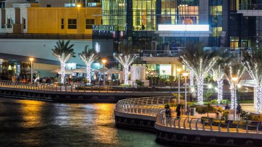 Yürüyen insanlarla dolaş ve Dubai Marina Kanalı üzerinde trafiği olan köprü gece vakti, BAE. Palmiyeler, tekneler ve kulelerle rıhtımın manzarası. Yapay kanallı bölge