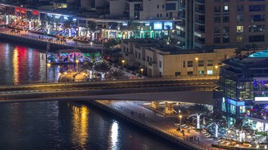 Dubai Marina 'daki su kanalı ve gezinti güvertesi gece vakti ufuk çizgisinde. Işıklı ve aydınlatmalı kuleler. Köprüde trafiği olan yüzen yatlar ve tekneler
