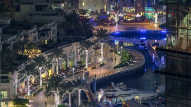 Dubai Marina 'daki gezinti güvertesi ve kanalı. Gece vakti, Birleşik Arap Emirlikleri civarında palmiyeler ve yatlarla. Yürüyüş yolunda restoranları ve kafeleri olan hava manzaralı.