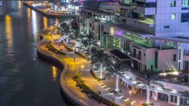 Dubai Marina 'daki rıhtım ve gezinti güvertesi gece vakti ufuk çizgisinde. Aydınlatma ve aydınlatma olan restoranlar. Gökdelenlerin yakınında trafik olan yüzen yatlar ve tekneler.