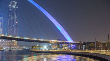 Dubai Su Kanalı üzerindeki fütürist yaya köprüsü gece saatlerinde aydınlandı, BAE. Arka planda deniz manzarası olan bir şehir. Gezinti güvertesinden göster
