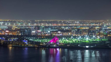 Şehirdeki golf sahasında binaların ışıkları ve aydınlatılmış yollarla gece vakti Dubai Deresi yakınlarındaki havadan zaman dilimi. Havalimanı arka planda, Dubai, Birleşik Arap Emirlikleri