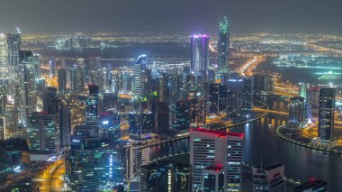 Dubai gece zaman atlamalı iş bölümü kuleleri panoramik havadan görünümü. Yapım aşamasındaki bazı ışıklı gökdelenlerin, kanalın ve yeni kulelerin çatı görünümü.