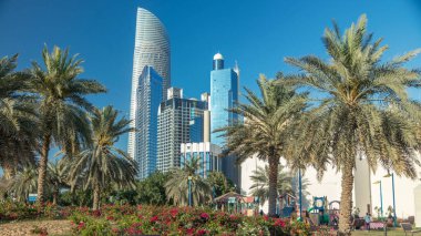 Abu Dabi 'deki sahil şeridi boyunca uzanan Corniche Bulvarı plajı parkında arka planda gökdelenler var. Aile parkından görüntü. Avuçlar yana. Güneşli bir günde mavi gökyüzü