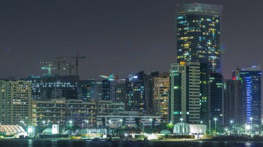 Abu Dabi Skyline 'ın panoramik görüntüsü ve gece vakti deniz manzarası, Birleşik Arap Emirlikleri. Aydınlatılmış gökdelenleri ve şantiyesi olan Corniche manzarası