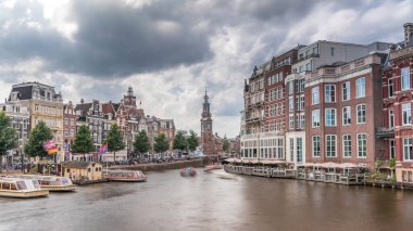 Amstel Nehri, rıhtım ve Munttoren kulesinin yer aldığı Amsterdam şehir merkezinin panoramik zaman atlaması. Tekneler bulutlu gökyüzünün altında ileri geri yüzerler. Köprüden manzaralı şehir manzarası. Hollanda