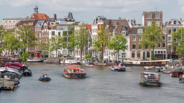 Güneşli bir günde Amsterdam 'ın şehir merkezindeki işlek bir nehrin zamanı. Rıhtımı çevreleyen yeşil ağaçlı tekneler trafiği. Arka planda tipik tarihi binalar. Amsterdam, Hollanda