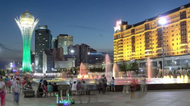Bayterek Kulesi ve çeşme gösterisi günden geceye geçiş zamanı. İnsanlar gün batımından sonra etrafta dolaşıyor. Arka planda gökdelenler var. Astana 'daki anıt ve gözlem kulesi. Nur-Sultan şehri, Kazakistan