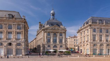 Place de la Bourse, Fransa 'nın Bordeaux şehrinde, bulutlu mavi gökyüzü altında klasik mimari ile çevrili restoranları olan bir yer. Meydanda ve ana caddelerde yürüyen insanlar
