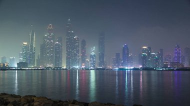 Dubai Marina night skyline Timelapse with fog as seen from Palm Jumeirah, Dubai, UAE. Illuminated skyscrapers reflected on water