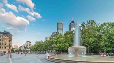 Frankfurt, Almanya merkezindeki Opera Meydanı (Opera Meydanı) zaman atlaması, arka planda gökdelenler ve kulelerle çevrili ve akşam gökyüzü altında ağaçlarla çevrili bir çeşme ve şehir hayatı sergiliyor.