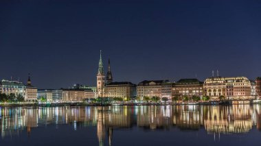 Binnenalster 'in Hamburg, Almanya' daki Belediye Binası ve Nikolai Kilisesi 'ndeki gece programı. Aydınlatılmış tarihi binalar yıldızlı bir gökyüzünün altındaki Alster Gölü sularına yansıyor. Manzaralı şehir manzarası