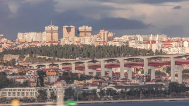 Renkli evler, Amoreiras alışveriş merkezi kuleleri ve 25 Nisan Köprüsü 'nün yer aldığı Lizbon siluetinin hava zaman çizelgesi. Önünde yeşil ağaçlar ve Prazeres Mezarlığı bulunan tarihi bölge