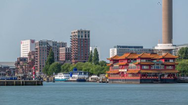 Euromast gözlem kulesi yakınlarındaki Rotterdam şehir manzarası zaman çizelgesi ve Nieuwe Maas Nehri 'ndeki bir turist teknesi. Rıhtımda modern gökdelenler, yüzen bir Çin restoranı ve manzaralı bir liman manzarası yer alıyor.