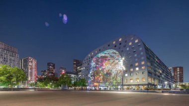 Rotterdamse Markthal 'ın dış görünüşünün geceden güne geçişi. Dükkanları ve restoranları olan ikonik bir bina. Meydanda gezinen insanlar. Gökdelenler ve arka planda canlı bir şehir manzarası